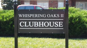 Sign for Whispering Oaks II Clubhouse in front of greenery and a vehicle.