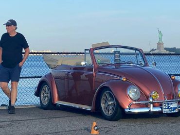 Vintage convertible car parked by the waterfront with a man standing nearby and the Statue of Liberty in the background.