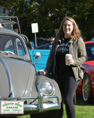 Woman smiling near a vintage car holding a Dunkin' cup.