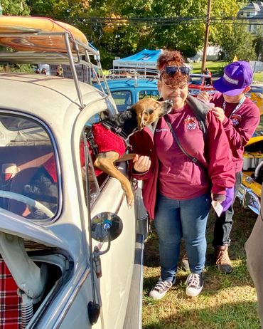 Dog affectionately licking woman's face near vintage car at outdoor event.