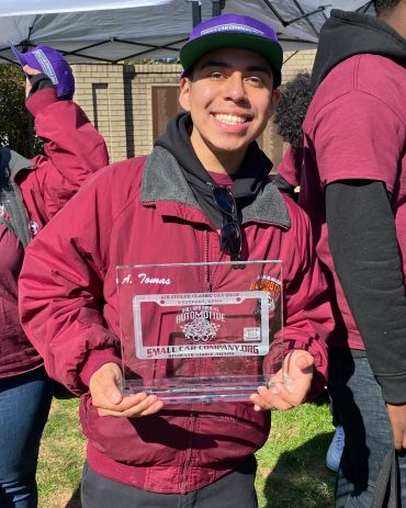 Smiling young man holding an automotive award plaque outdoors.