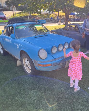 A little girl in a strawberry dress points at a blue vintage car with extra lights on the hood.
