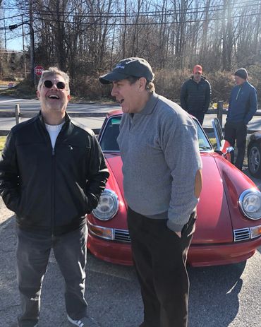 Two men laughing beside a red vintage car on a sunny day.