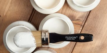 Paintbrush dipped in white paint with open paint containers on wooden surface.