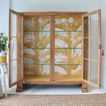 Wooden display cabinet with glass shelves and tropical wallpaper backdrop.