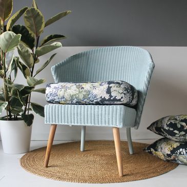 Light blue corduroy chair with floral cushion and wooden legs on a round rug.