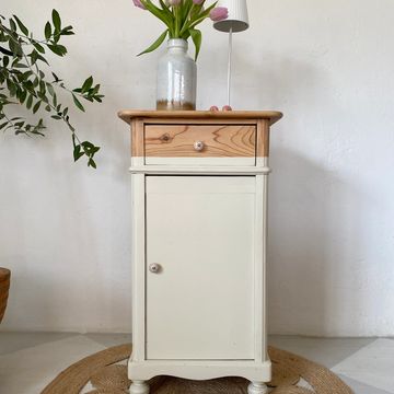 A small wooden cabinet with a drawer and a door, decorated with a vase of flowers and a lamp.