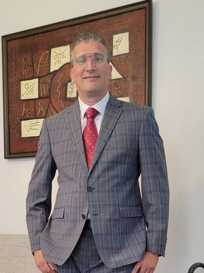 Man in a gray plaid suit with a red tie standing indoors.