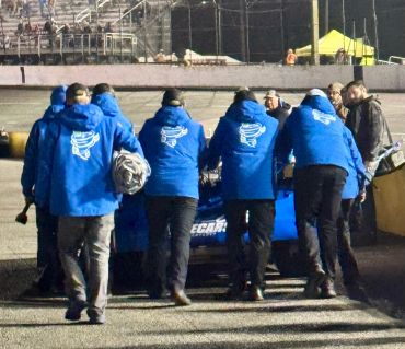 A team in blue jackets pushes a race car on a track at night.