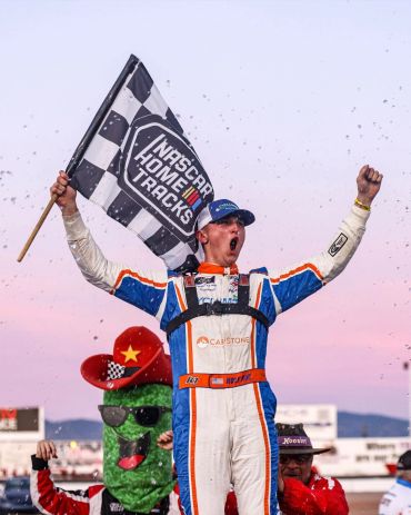 Race car driver celebrates victory holding a NASCAR Home Tracks flag.