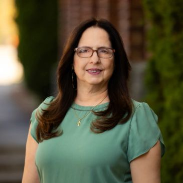 Woman with glasses wearing a green top and cross necklace.
