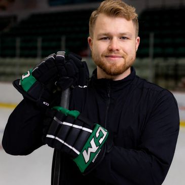 Smiling hockey player in black jacket holding a hockey stick on ice rink.