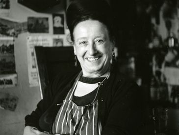 Smiling woman wearing an apron in a cozy, cluttered room.
