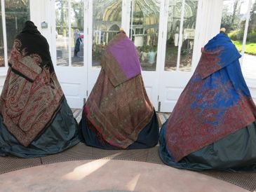 Three figures draped in patterned shawls standing indoors near glass doors.