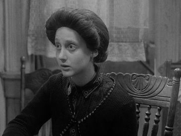 A woman with a surprised expression sits at a wooden chair indoors.