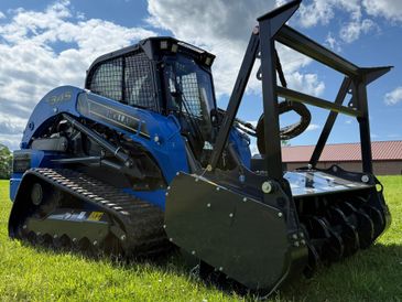 Blue tracked skid steer with a heavy-duty attachment on grass.