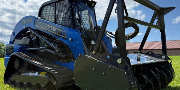 Blue New Holland C345 compact track loader with forestry mulcher attachment on grass.