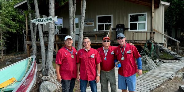 Four men in red shirts posing outdoors near a cabin.