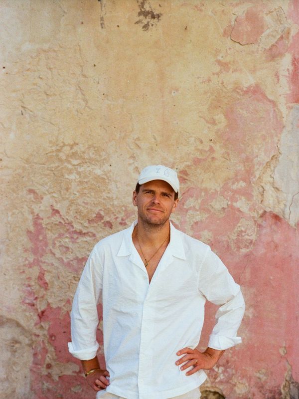 Man in white shirt and cap standing against a textured pink wall.