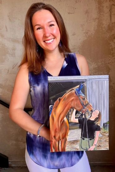 Woman smiling while holding a painting of herself with a horse.
