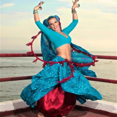 A woman joyfully dances in vibrant blue traditional attire by the sea.