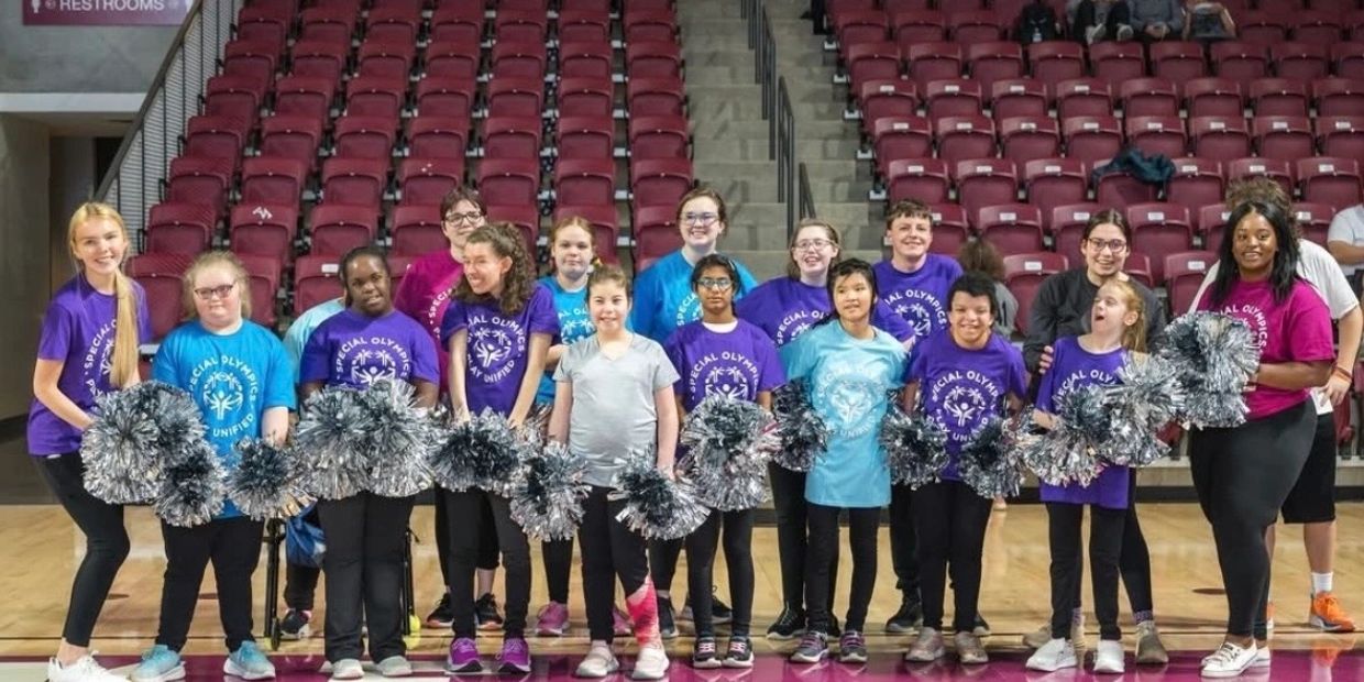 A diverse group of young cheerleaders holding silver pom-poms in a gymnasium.