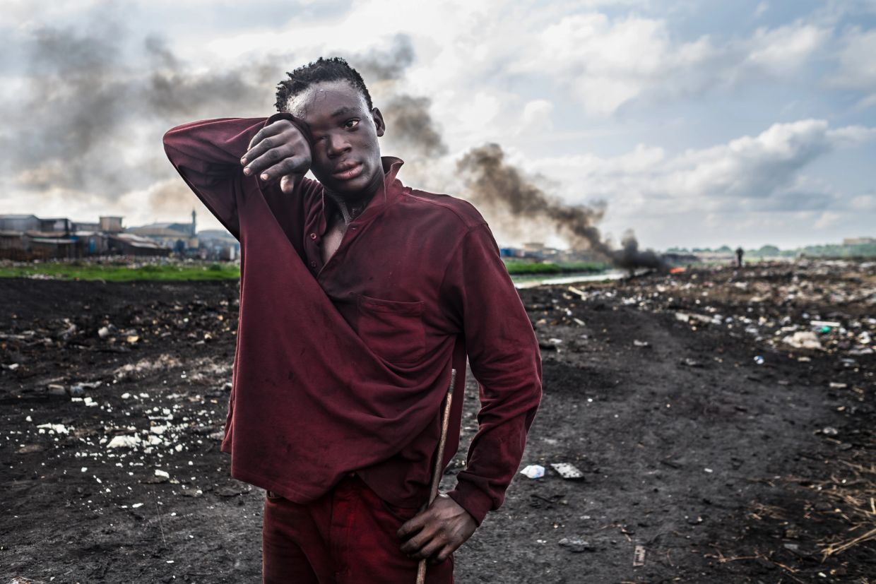 Young man in maroon clothes wiping sweat in a smoky, trash-filled area.