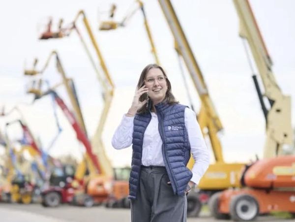Woman in a blue vest talking on the phone at a construction site with cranes.