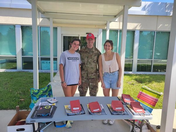 Two women and a soldier stand behind a table with books and pamphlets outdoors.
