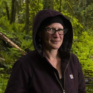 White woman wearing a black hoodie and glasses smiling, with forest and ferns in the background