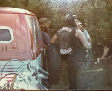 Group of people standing near a vintage van and motorcycle in a wooded area.