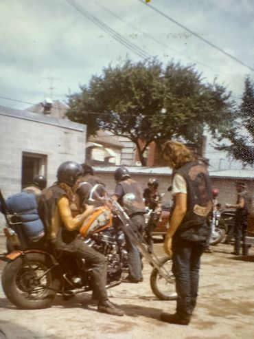 Bikers with helmets and vests gathered around motorcycles in a sunny outdoor setting.