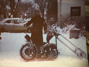 Man standing next to a custom chopper motorcycle in a snowy setting.