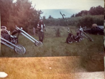 Vintage photo of chopper motorcycles and a person in a grassy field.