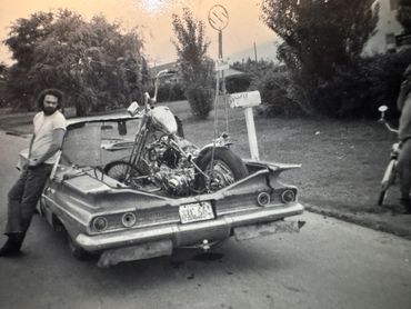 Man leaning on a modified car carrying a motorcycle in its bed.