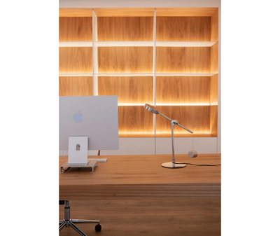 Minimalist wooden desk with an Apple iMac and sleek desk lamp in front of illuminated wooden shelves.
