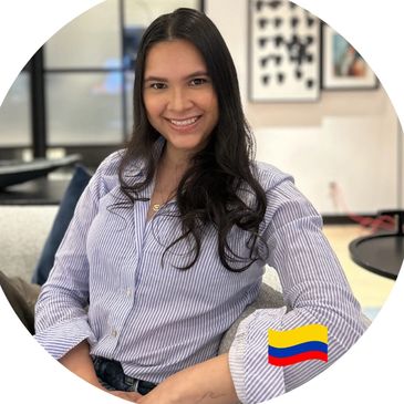Smiling woman sitting indoors, dressed in a striped shirt with a Colombian flag icon.