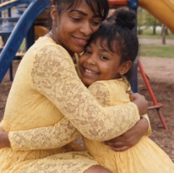 Mother and daughter in matching yellow lace dresses sharing a warm hug outdoors.