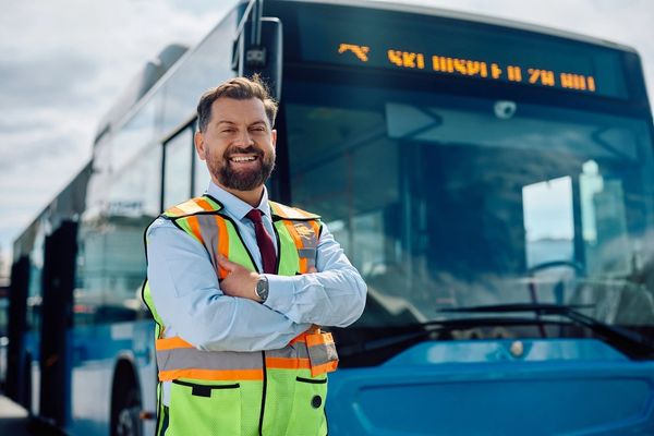 A smiling, bearded bus driver in a light blue shirt, red tie, and a reflective vest, arms crossed