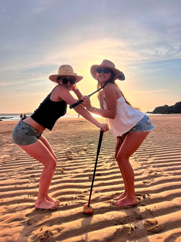 Two women posing with a golf putter on the beach at sunset.