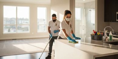 Two masked cleaners sanitizing a modern kitchen and living room area.