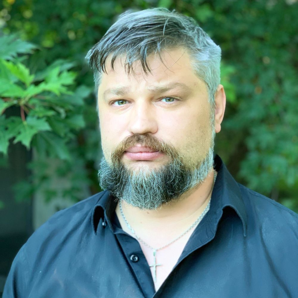 Man with gray beard and hair wearing a black shirt and cross necklace outdoors.