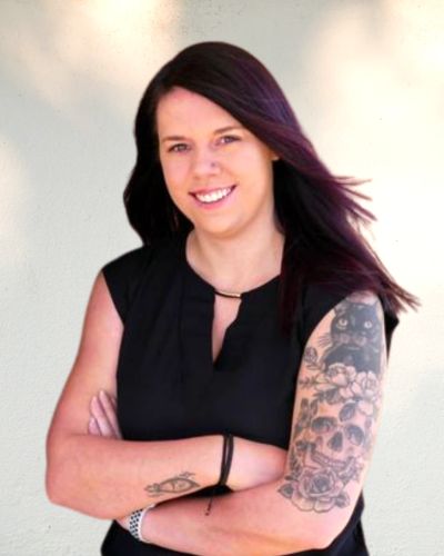 Smiling woman with dark hair and detailed arm tattoos, wearing a black sleeveless top.