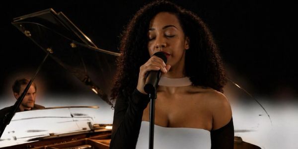 A woman sings passionately into a microphone with a pianist in the background.