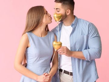 Couple sharing a kiss behind heart-shaped balloons on a pink background.
