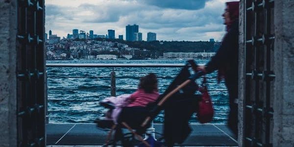 una mamma che spinge una bambina nel passeggino in riva al fiume con lo sfondo di uno skyline
