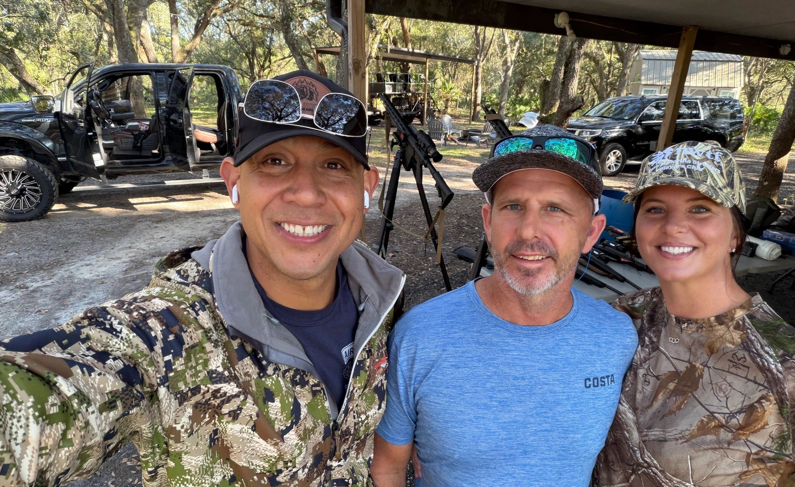 Three people smiling outdoors with hunting gear and trucks nearby.