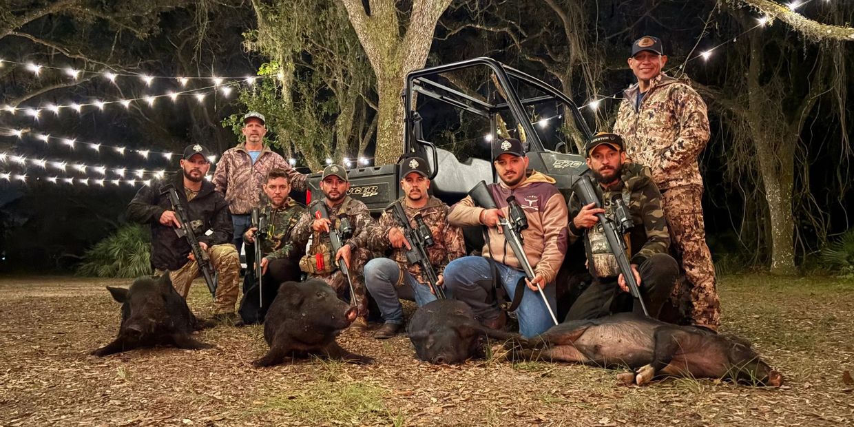 Group of hunters posing with rifles and wild boars at night under string lights.