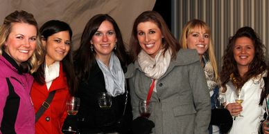 Six women smiling and holding drinks at a social gathering indoors.