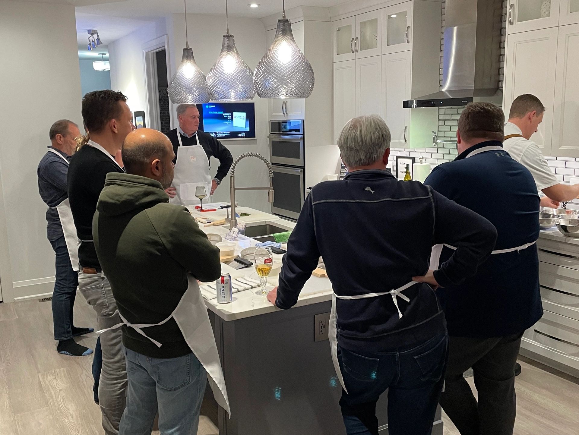 Group of men in aprons gathered around a kitchen island during a hands-on cooking class.